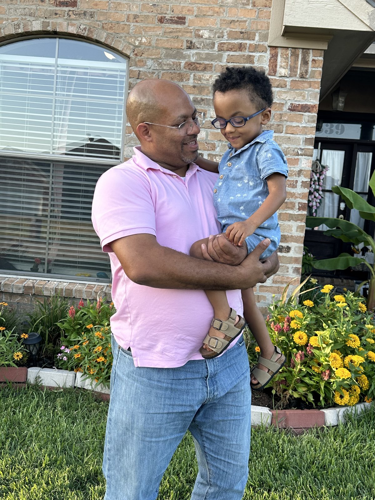 A smiling father holds his young son in glasses in front of their home.